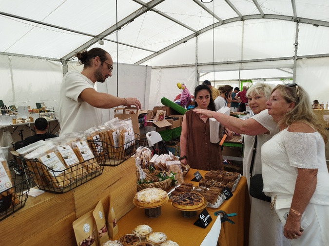 Elisabeth tend le bras pour récupérer les pâtisseries qu'elle vient d'acheter à Pauline (au centre), sa pâtissière favorite.